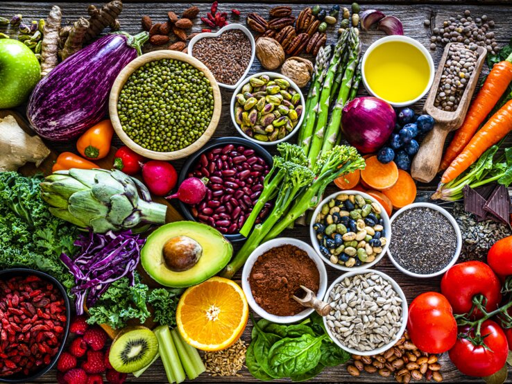 Ein reichhaltiges und farbenfrohes Arrangement von Superfoods und gesunden Lebensmitteln auf einem rustikalen Holztisch. Zu den sichtbaren Lebensmitteln gehören verschiedene frische Gemüsesorten wie Auberginen, Tomaten und grünes Blattgemüse, Obst wie Äpfel und Beeren, Nüsse, Samen, Hülsenfrüchte und Gewürze. Das Bild strahlt Frische und Vielfalt aus und betont eine nährstoffreiche Ernährung.
