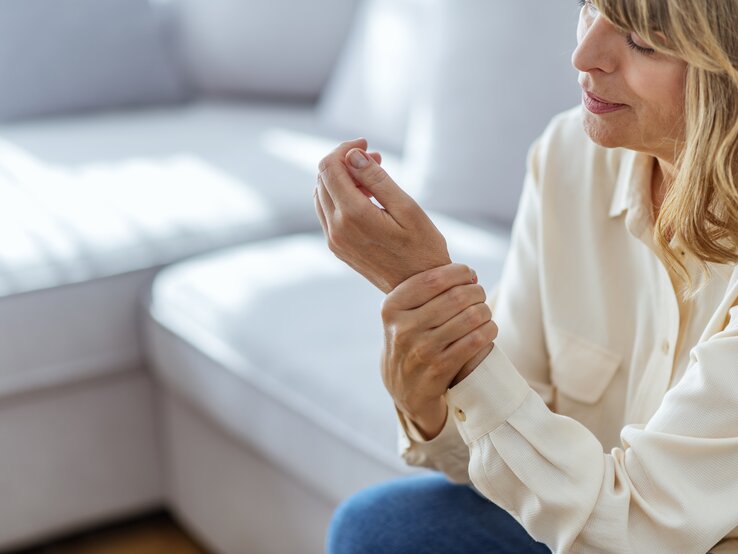 Eine Frau mittleren Alters in einem hellen, lässigen Hemd sitzt und betrachtet besorgt ihr schmerzendes Handgelenk. Sie hält mit ihrer anderen Hand das Handgelenk, was auf Unbehagen oder Schmerzen hindeutet. Ihr Gesichtsausdruck spiegelt Sorge und Unbehagen wider. Sie befindet sich in einem hellen Raum mit weicher Einrichtung im Hintergrund, was auf eine häusliche oder möglicherweise medizinische Umgebung hindeutet.