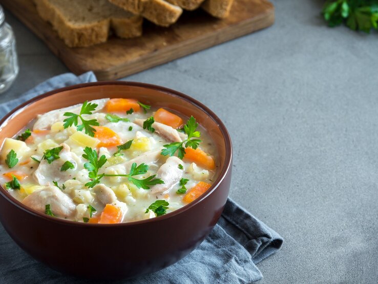 Eine braune Schale mit der Hühnersuppe auf grauem Untergrund. Im Hintergrund Petersilie, Brotscheiben und Pfeffer.