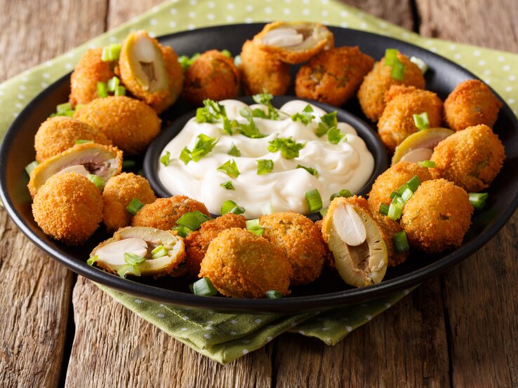 Auf einem schwarzen Teller liegen gebratene Oliven mit Mandeln. In der Mitte des Tellers ist eine weiße Soße.