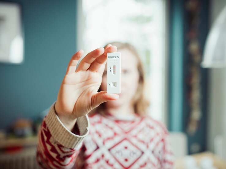 Eine Frau mittleren Alters, die einen rot-weiß-gemusterten Pullover trägt, hält einen positiven Corona-Test in die Kamera, der anzeigt, dass sie die Corona-Mutation Priola hat.