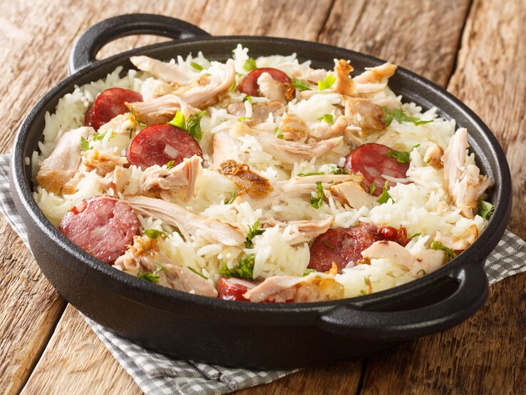 One-Pot-Reis-Huhn One Pot Reis mit Huhn und gebratener Wurst in Gusspfanne auf Stoffserviette auf Holztisch. Aufnahme aus der Draufsicht.