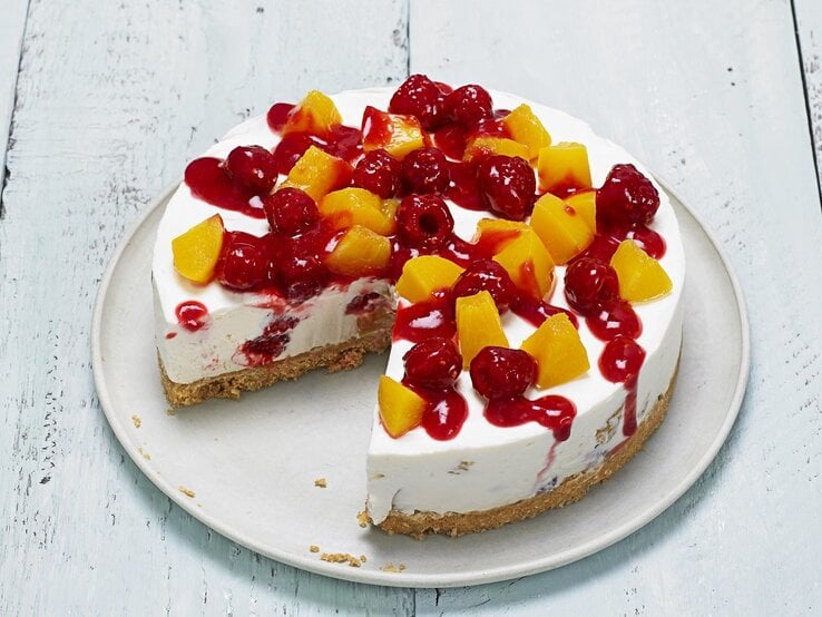 Unser Pfirsich-Melba-Käsekuchen bringt Abwechslung in die Winter-Backstube. So fruchtig! Auf einem Teller liegt ein Pfirsich-Melba-Käsekuchen. Er ist mit Himbeersoße überzogen.