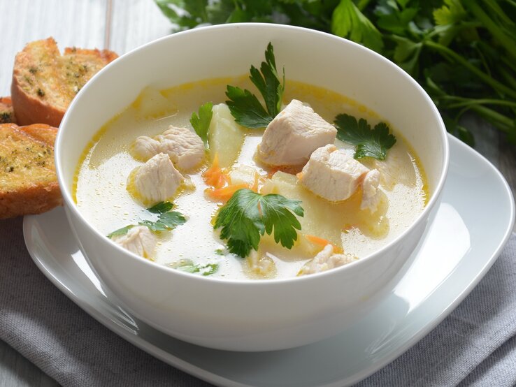 Eine weiße Suppenschale mit der Hühnerfrikassee-Suppe auf einem hellen Holztisch seitlich von schräg-oben fotografiert. Daneben noch ein Bund frische Petersilie auf der linken Seite.