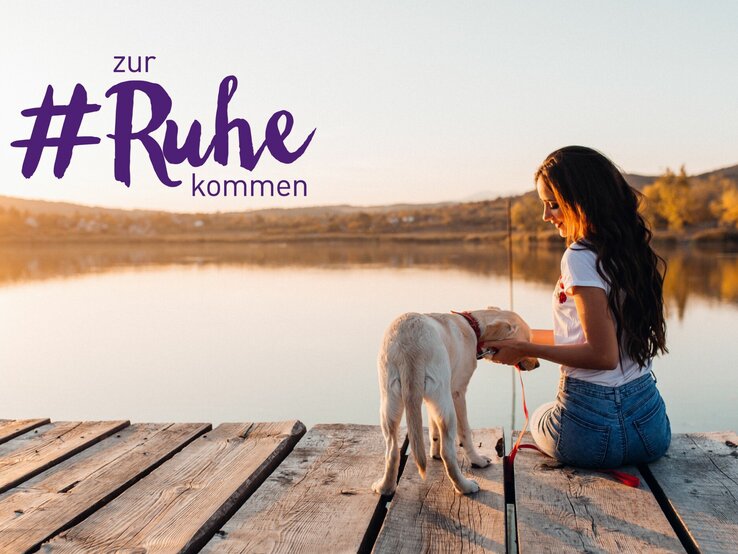 Eine Frau sitzt entspannt auf einem Holzsteg am Ufer eines Sees und streichelt liebevoll einen weißen Hund, der neben ihr steht. Sie trägt ein weißes T-Shirt und Jeans, und ihre langen dunklen Haare fallen über ihre Schultern. Die Szene ist in warmes, weiches Abendlicht getaucht, das eine friedliche und ruhige Atmosphäre schafft. Im Hintergrund sind sanfte Hügel und der ruhige See zu sehen. Über der Szene steht der Schriftzug "#zur Ruhe kommen" in großer lila Schrift, was auf einen Moment der Entspannung und des Einklangs mit der Natur hindeutet.