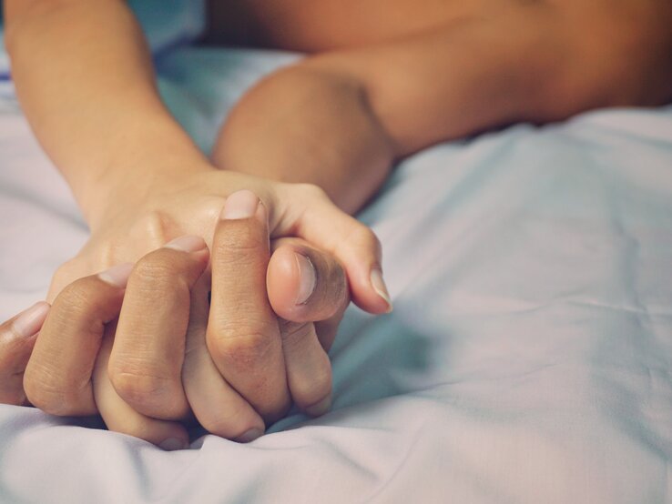 Zwei Hände, die sich fest auf einer blauen Bettdecke halten, wobei eine Person im Hintergrund undeutlich zu sehen ist, was Intimität und Nähe suggeriert.