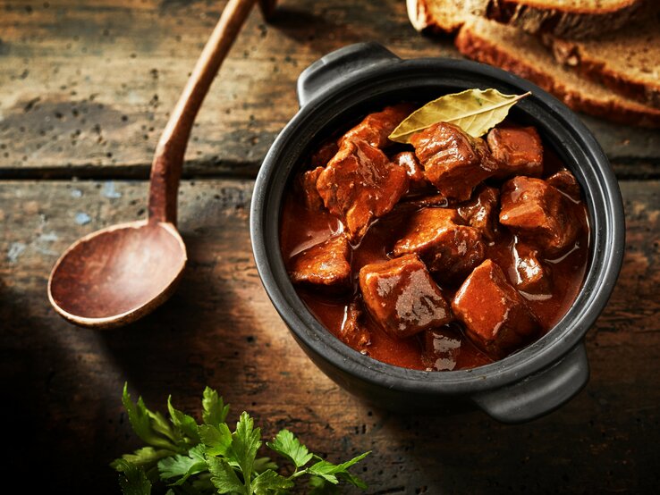 Gulasch in einem kleinen schwarzen Tontopf auf einem rustikalen Holztisch mit einem Holzkochlöffel und Petersilie.