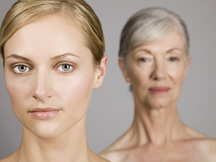 Zwei Frauen in einem Bild, die jüngere im Vordergrund mit Blick in die Kamera und die ältere im unscharfen Hintergrund, symbolisieren verschiedene Generationen oder den Alterungsprozess.