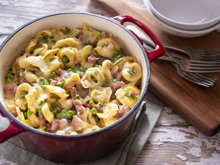 Tortellini mit Schinken und Erbsen in Sahnesoße, angerichtet in einem Topf. Daneben finden sich Holzbrett, Schalen und Gabeln. 