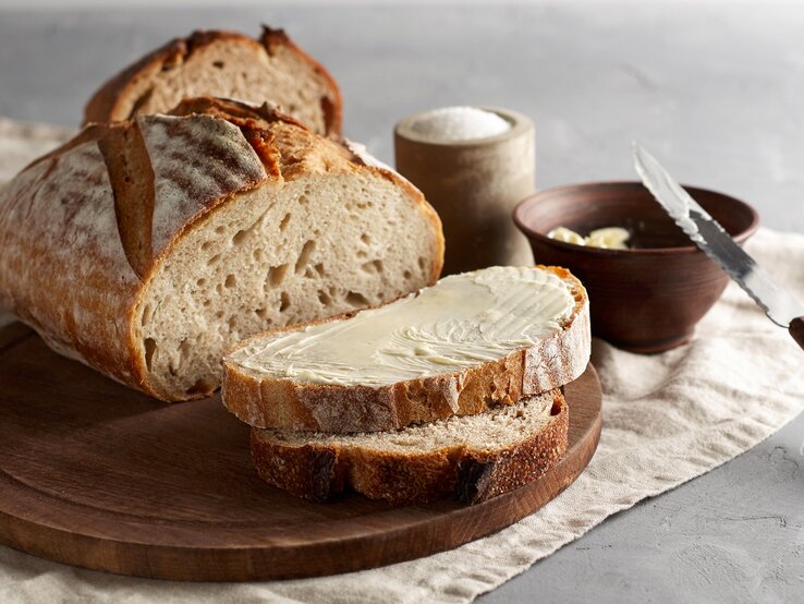 Ein angeschnittener Laib "Friss dich dumm"-Brot auf einem Brett, zwei Scheiben liegen davor, eine Scheibe mit Butter bestrichen, daneben eine Schale Butter mit einem Messer und eine Schale Salz.