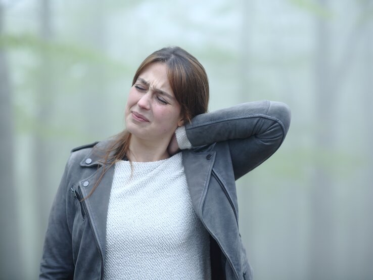 Eine junge Frau mit schmerzverzerrtem Gesicht hält sich den Nacken, als ob sie starke Schmerzen hätte. Sie trägt eine graue Lederjacke und einen hellen Strickpullover. Der Hintergrund ist verschwommen, mit sanften grünen Farbtönen, was auf eine Außenaufnahme hindeutet.