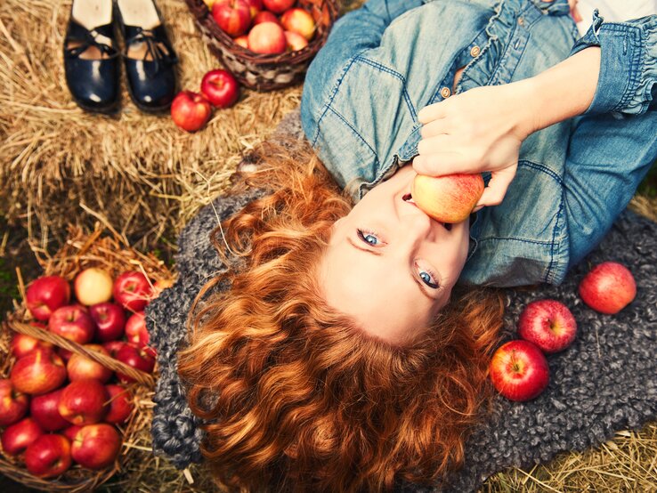 Das Bild zeigt eine junge Frau mit lockigem, rotem Haar, die entspannt auf dem Rücken liegt und direkt in die Kamera blickt. Sie hält einen Apfel in ihrer Hand nahe ihrem Gesicht, was eine spielerische und naturnahe Atmosphäre vermittelt. Sie ist auf einer grauen Decke platziert, die auf einer Wiese ausgebreitet ist, und um sie herum sind frische rote Äpfel verstreut. Ein Korb voller Äpfel ist neben ihr zu sehen, ebenso wie ihre Schuhe, die sorgfältig neben einem Strohballen abgestellt sind. Das Bild strahlt eine herbstliche Stimmung aus und könnte in einem ländlichen oder Apfelernte-Kontext stehen.