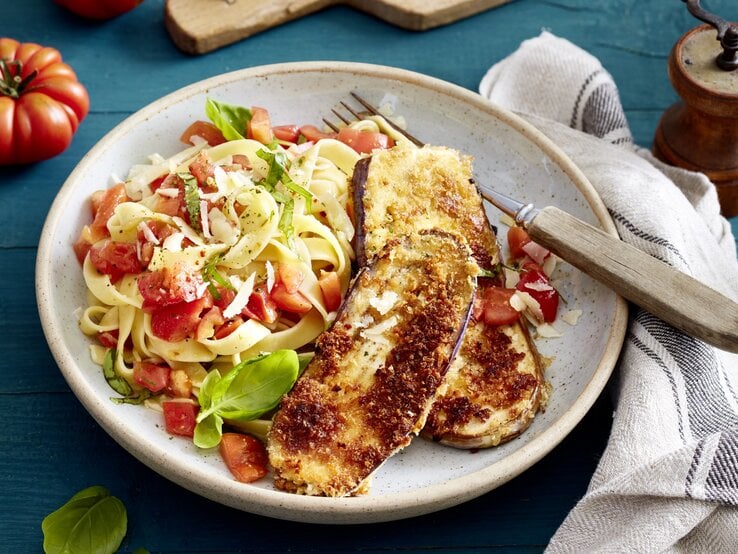 Auberginen-Schnitzel mit Bruschetta-Nudeln und Gabel auf einem hellen Teller. Drum herum Basilikum, Tomaten und ein Küchentuch, alles auf blauem Untergrund. 
