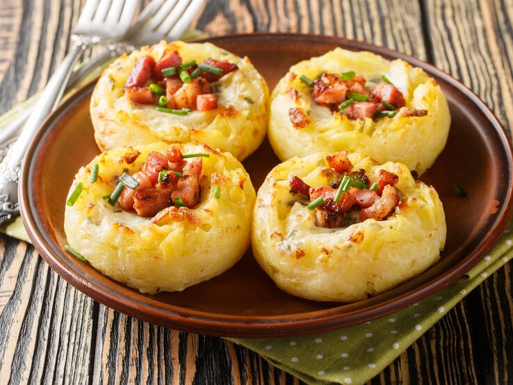 Ein brauner runder Teller mit vier Speck-Kartoffelnestern auf einem braunen Holztisch.