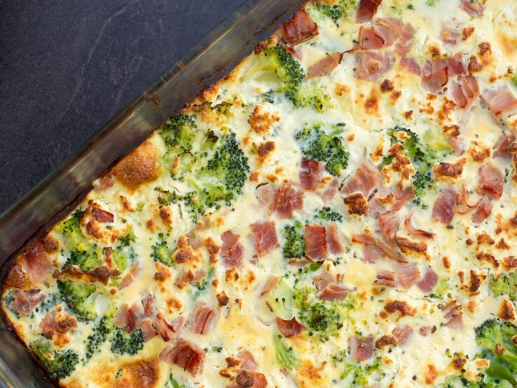 Gläserne Auflaufform gefüllt mit übebackenem Brokkoli-Schinken-Gratin auf dunklem Untergrund. 
