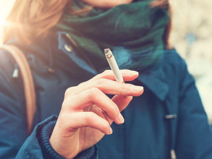 Eine junge Frau hält eine brennende Zigarette zwischen Zeige- und Mittelfinger. An den Stellen, an denen die Zigarette die Haut berührt, werden sich mit der Zeit gelbe Flecken bilden. Wie wird man sie los?