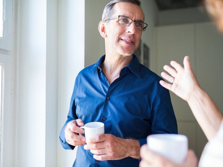 Ein hört zu, was seine Frau ihm sagt und steht dabei mit einer Tasse am Fenster. | © iStock/alvarez