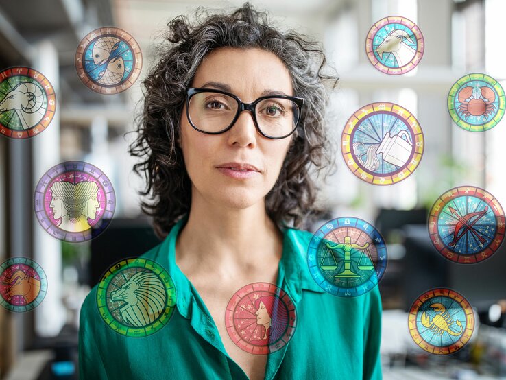 Eine Frau mit lockigen grauen Haaren und Brille steht in einem Büro. Die astrologischen Symbole der 12 Sternzeichen umgeben sie. | © iStock [M]