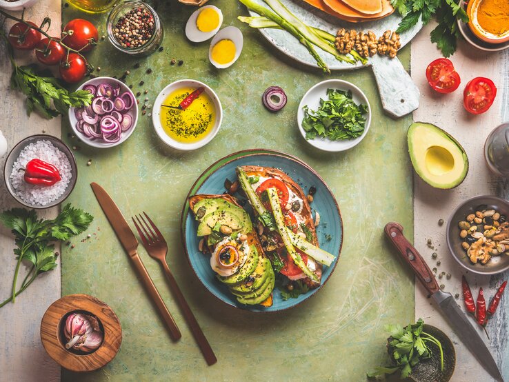 Ein Bild von einem reichhaltigen, farbenfrohen vegetarischen Mahl auf einem rustikalen Tisch. Im Zentrum befindet sich ein Teller mit einem offenen Avocado-Sandwich, belegt mit Tomaten, Zwiebeln und Gewürzen.
