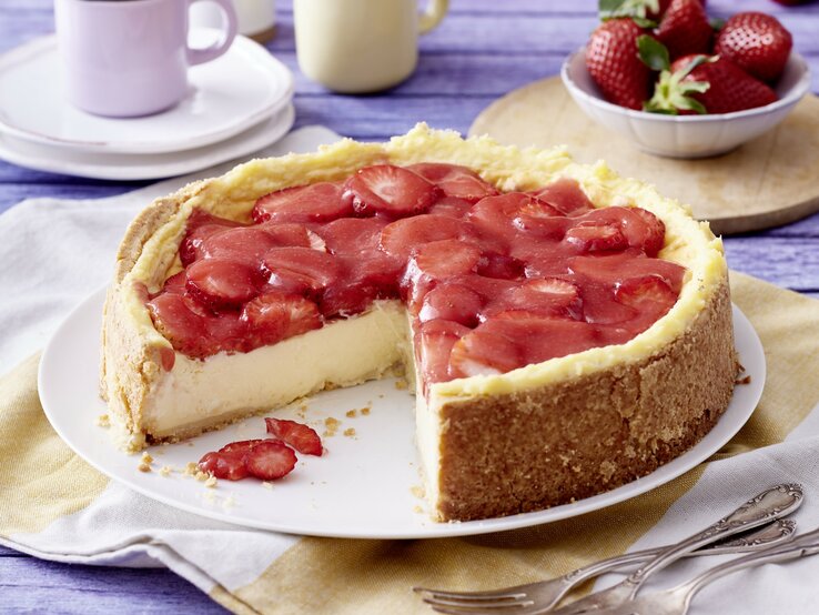 Angeschnittener Erdbeer-Schmandkuchen auf weißem Teller, daneben Gabeln, Geschirr und Erdbeeren auf Küchentüchern mit blauem Holzuntergrund. 