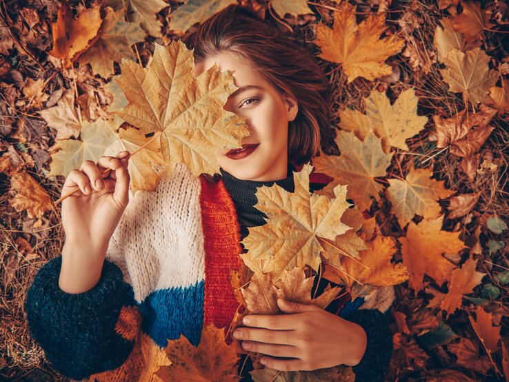 Eine junge Frau liegt auf einem Bett aus bunten Herbstblättern. Sie hält ein großes, gelbes Ahornblatt vor ihrem rechten Auge, wodurch nur die linke Gesichtshälfte sichtbar ist. Sie trägt einen dunklen Lippenstift, der gut zum Herbst passt. Ihr Haar, das teilweise unter den Blättern verborgen ist, hat eine brünette Färbung. Sie trägt eine gestrickte Jacke mit mehrfarbigen Streifen in Weiß, Orange, Rot und Blau.