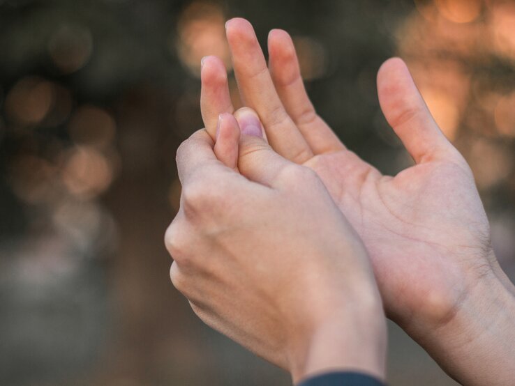 Zwei Hände sind zu sehen. Die rechte Hand massiert die Innenseite des linken Ringfingers, der Schmerzen zu verursachen scheint, was beim paroxysmale Fingerhämatom üblich ist. Der Hintergrund ist unscharf.