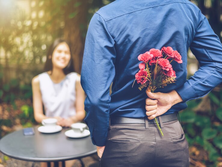 Ein junger Mann hält einen Blumenstrauß hinter seinem Rücken, vor ihm sitzt eine junge Frau und lächelt ihn an
