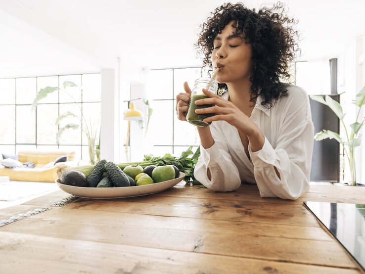 Eine Frau mit lockigem Haar genießt einen grünen Smoothie, während sie an einem Holztisch sitzt. Vor ihr liegt ein Teller mit frischen grünen Früchten und Gemüse wie Avocados und Limetten. Sie trägt eine weiße Bluse und der Raum ist hell und freundlich, mit einem großen Fenster und Pflanzen im Hintergrund, was eine gesunde und entspannte Atmosphäre schafft.