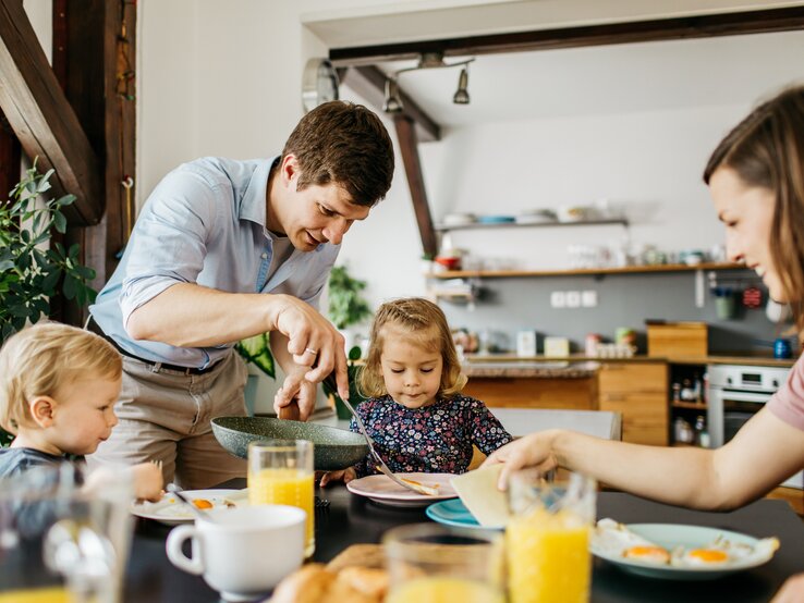 Eine glückliche Familie beim Frühstück: Ein Mann in einem hellblauen Hemd beugt sich über, um seiner kleinen Tochter zu helfen, die konzentriert auf ihren Teller blickt. Ein weiteres Kleinkind sitzt daneben und ist auf sein Essen fokussiert. Eine Frau sitzt gegenüber, reicht etwas hinüber und lächelt dabei. Der Tisch ist mit einem gesunden Frühstück mit Orangensaft, Spiegeleiern und Brötchen gedeckt. Sie befinden sich in einer hellen Küche mit moderner Einrichtung und Pflanzen, die für eine wohnliche Atmosphäre sorgen.
