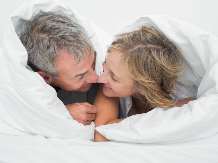 Ein Paar liegt nebeneinander im Bett und schaut sich lächelnd an, die Bettdecke ist über beide Köpfe gezogen