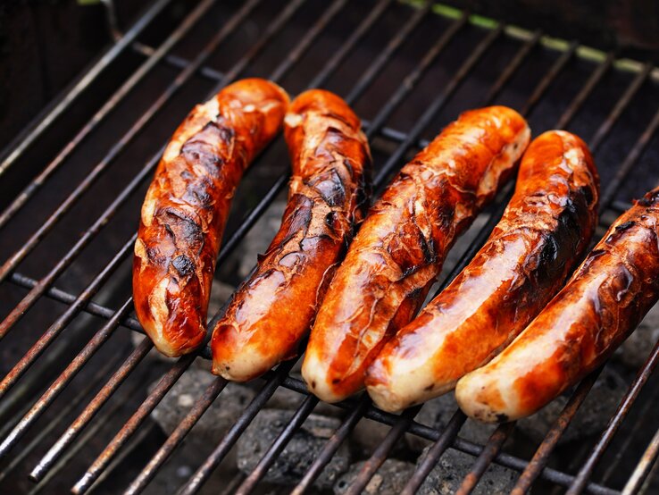 Ran an die Wurst: Die weltberühmte Thüringer Rostbratwurst wird traditionell über einem Holzkohlenfeuer gegrillt! Draufsicht: Fünf Thüringer Rostbratwürste liegen auf einem Holzkohlegroll.