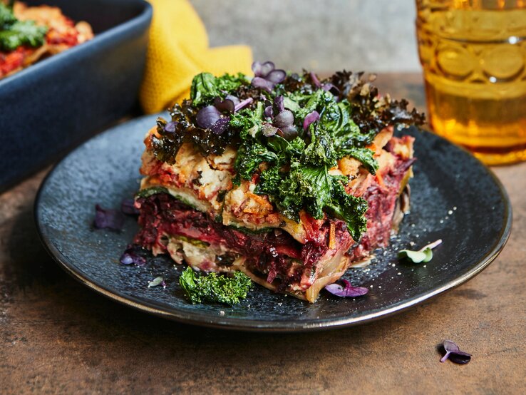 Eine Portion bunte Winterlasagne mit Grünkohl und rote Bete steht auf einem dunklen Teller auf einem Holztisch. Die Lasagne ist mit bunter Kresse angerichtet. Im Hintergrund steht die Ofenform mit der restlichen Lasagne und ein Glas.