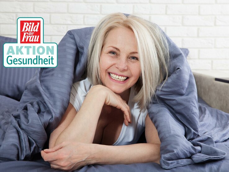 Eine fröhliche Frau mittleren Alters mit strahlendem Lächeln liegt entspannt im Bett, umhüllt von einer grauen Bettdecke. Sie hat kurzes, platinblondes Haar und trägt ein weißes Top, das Frische und Wohlgefühl ausstrahlt. Ihr Kopf ruht auf ihren verschränkten Armen, und ihre Augen sind vor Freude leicht geschlossen. Die weiße Ziegelwand im Hintergrund vermittelt ein modernes, wohnliches Ambiente. In der oberen linken Ecke ist das Logo "Bild der Frau AKTION Gesundheit" zu sehen.