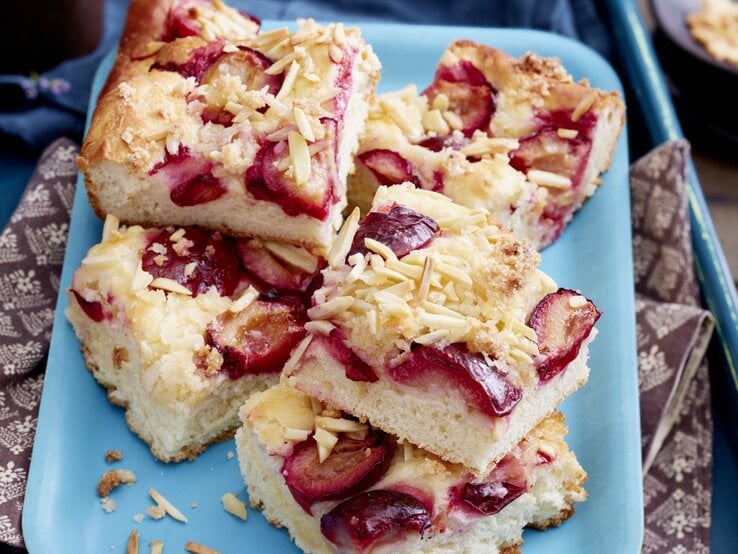 Pflaumen-Butterkuchen vom Blech, geschnitten und aufeinander auf einem blauen Teller gestapelt serviert. Die einzelnen Kuchenstücke mit Pflaumen und Mandelsplittern garniert. 