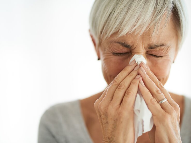 Eine Frau mit kurzen grauen Haaren benutzt ein Taschentuch, um ihre Nase zu putzen. Die Augen hat sie dabei geschlossen, was auf eine Erkältung oder eine Allergie hinweisen könnte. Die Frau steht vor einem unscharfen, hellen Hintergrund.
