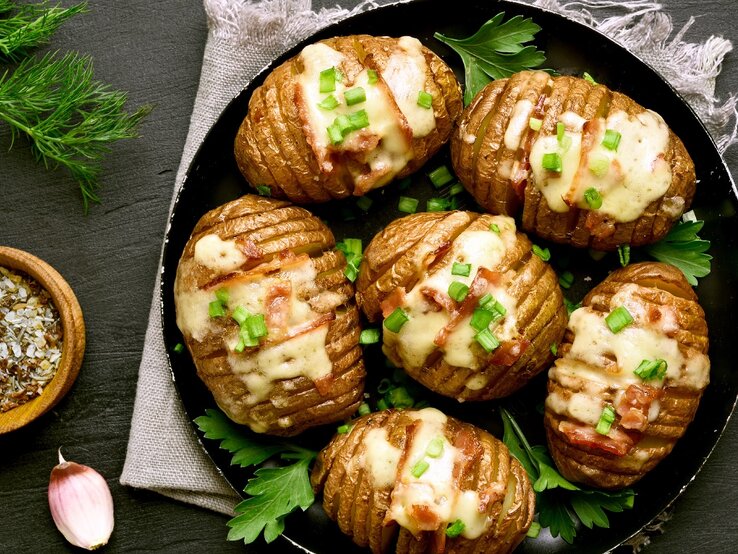 Eine Pfanne mit den Hasselback-Kartoffeln auf einer grauen Decke und einem Schiefer-Untergrund.