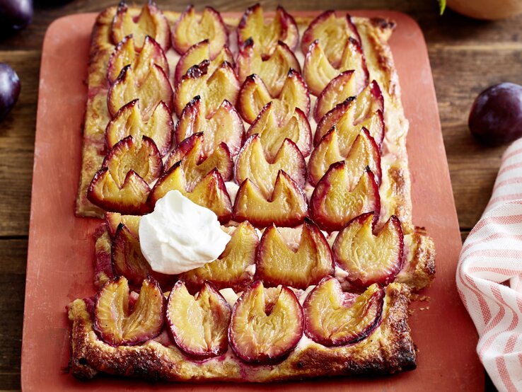 So eine fixe Pflaumen-Blätterteig-Tarte hat bisher wirklich jeden Tag bereichert. Angeschnittene Pflaumen-Blätterteig-Tarte mit Sahne auf einem blassroten Brett, im Hintergrund Pflaumen und ein Küchentuch, darunter Holz.