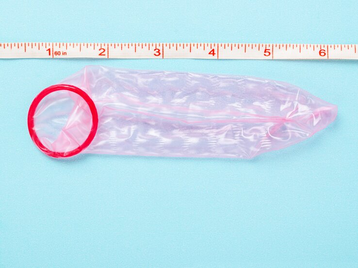 Ein rotes Kondom neben einem Maßband, das Zoll und Zentimeter anzeigt, auf einer hellblauen Oberfläche.