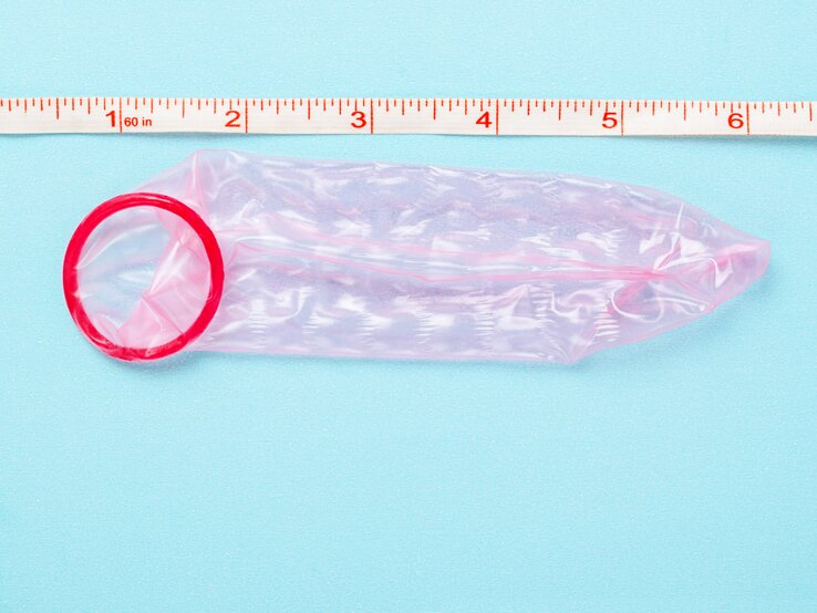 Kondomgröße berechnen Ein rotes Kondom neben einem Maßband, das Zoll und Zentimeter anzeigt, auf einer hellblauen Oberfläche.