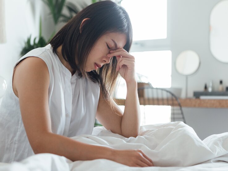  Das Bild zeigt eine junge Frau, die in einem hell erleuchteten, modern eingerichteten Schlafzimmer auf einem Bett sitzt. Sie trägt ein weißes Oberteil und hält mit geschlossenen Augen und einem schmerzverzerrten Gesichtsausdruck ihre Stirn, was darauf hindeutet, dass sie möglicherweise unter Kopfschmerzen oder Stress leidet. Die weiße Bettwäsche und das minimalistische Design des Zimmers sorgen für eine ruhige Atmosphäre, die im Gegensatz zu ihrem offensichtlichen Unbehagen steht.