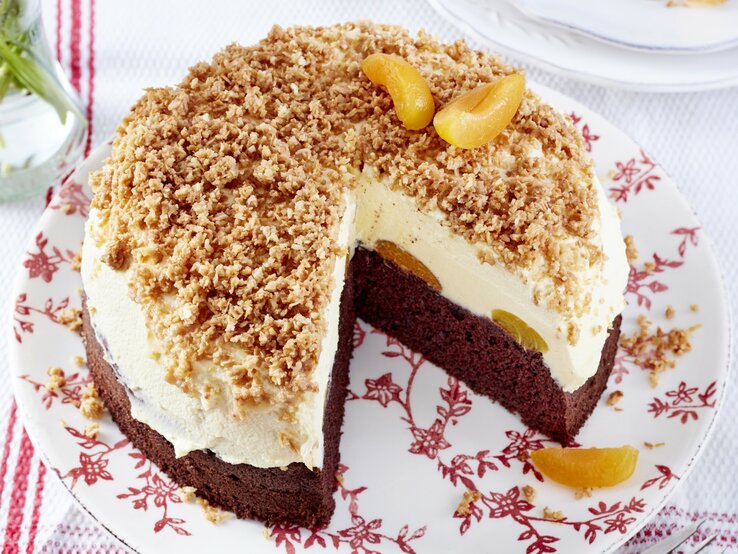 Sägespäne-Aprikosen-Torte mit einem Schokoladenboden, einer cremigen Füllung und einem knusprigen Dekor aus Kokosraspeln, angeschnitten auf einem blumigen Teller auf weißem Untergrund. 