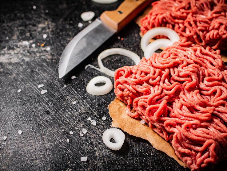 Draufsicht: Rohes Fleisch, das durch den Fleischwolf gedreht wurde liegt auf einem dunklen Untergrund. Daneben liegen rohe Zwiebelringe und ein Küchenmesser.