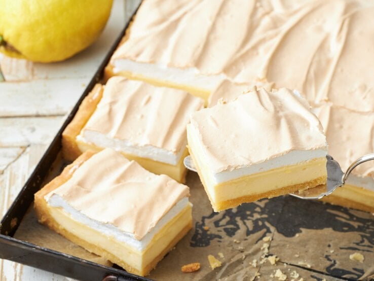 Zitronen-Schmand-Kuchen mit Baiser in einem dunklen Blech auf einem hellen Tisch mit frischen Zitronen. 