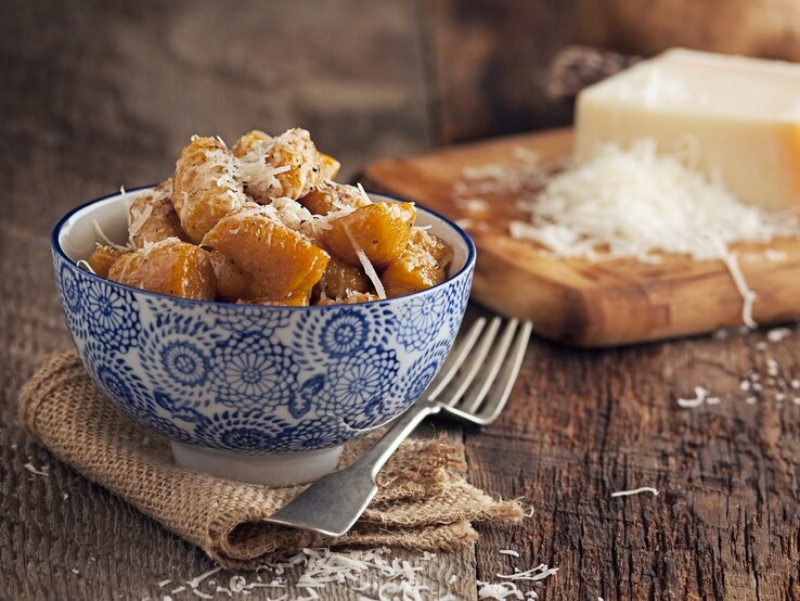 Eine Schale mit Süßkartoffel-Gnocchi, die mit Parmesan bestreut sind. Daneben eine Gabel, im Hintergrund ein Stück Parmesan.