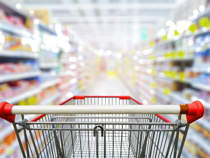 Ein leerer Einkaufswagen in einem Supermarktgang. Die Produkte sind unscharf zu erkennen.