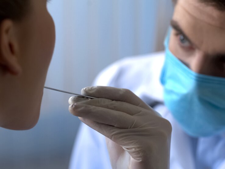 Ein Mediziner in blauer Schutzmaske führt mit Handschuhen einen Mundabstrich bei einer Patientin durch.
