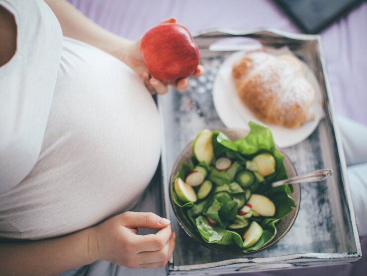 Das Bild zeigt eine schwangere Frau, die auf einem Bett sitzt. Ihr großer Babybauch ist unter einem engen, weißen Oberteil zu erkennen. Sie hält einen roten Apfel in ihrer rechten Hand und hat ein Holztablett auf ihren Knien, auf dem eine Schüssel mit grünem Salat und ein Teller mit einem Croissant liegen. Die Szene strahlt eine ruhige, häusliche Atmosphäre aus und betont die Bedeutung einer gesunden Ernährung während der Schwangerschaft.