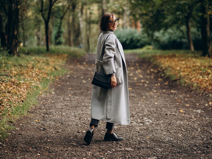 Eine Frau mittleren Alters steht im Herbst im Wald auf einem Waldweg. Sie hat braune Haare und trägt einen grauen Mantel.