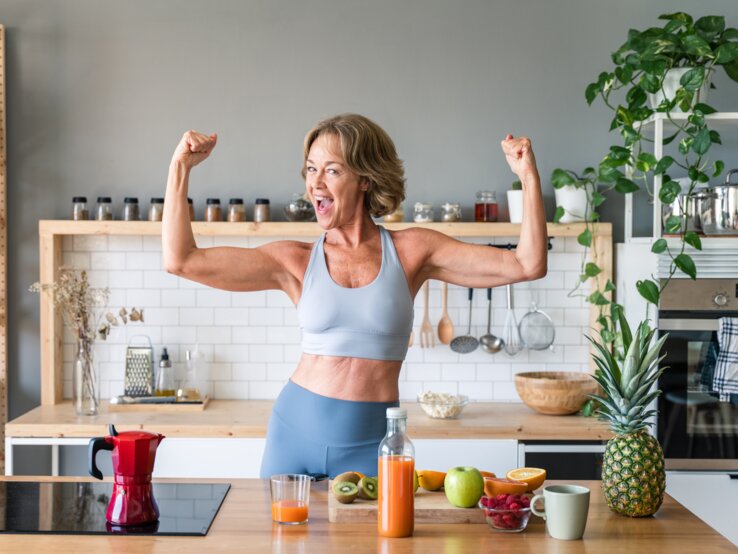 Eine ältere Frau, die Sportbekleidung trägt, steht in einer Küche und hat die Arme angewinkelt, sodass sie ihre definierten Arme präsentiert. Vor ihr auf dem Tisch liegt etwas Obst sowie eine Saftflasche.