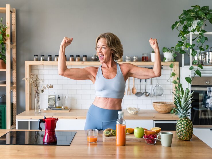 Eine ältere Frau, die Sportbekleidung trägt, steht in einer Küche und hat die Arme angewinkelt, sodass sie ihre definierten Arme präsentiert. Vor ihr auf dem Tisch liegt etwas Obst sowie eine Saftflasche.